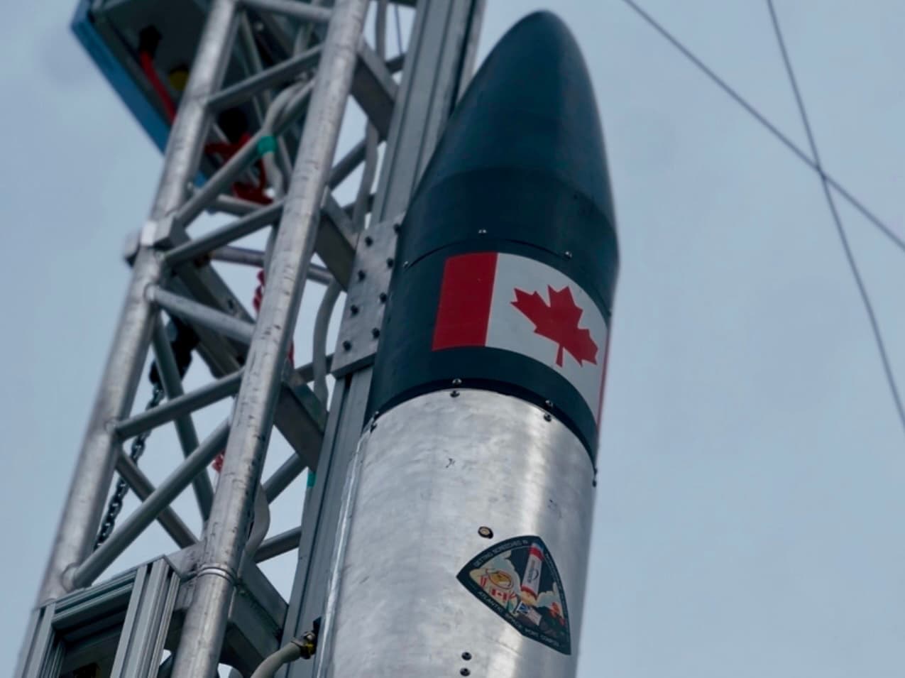 The nose cone of NordSpace's Taiga rocket on its launch tower, a Canadian flag on the fairing against an overcast sky